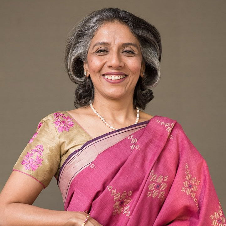 A smiling woman with gray hair styled in an updo, wearing a vibrant pink and gold floral-patterned sari with a beige embroidered blouse, posed against a solid taupe background.